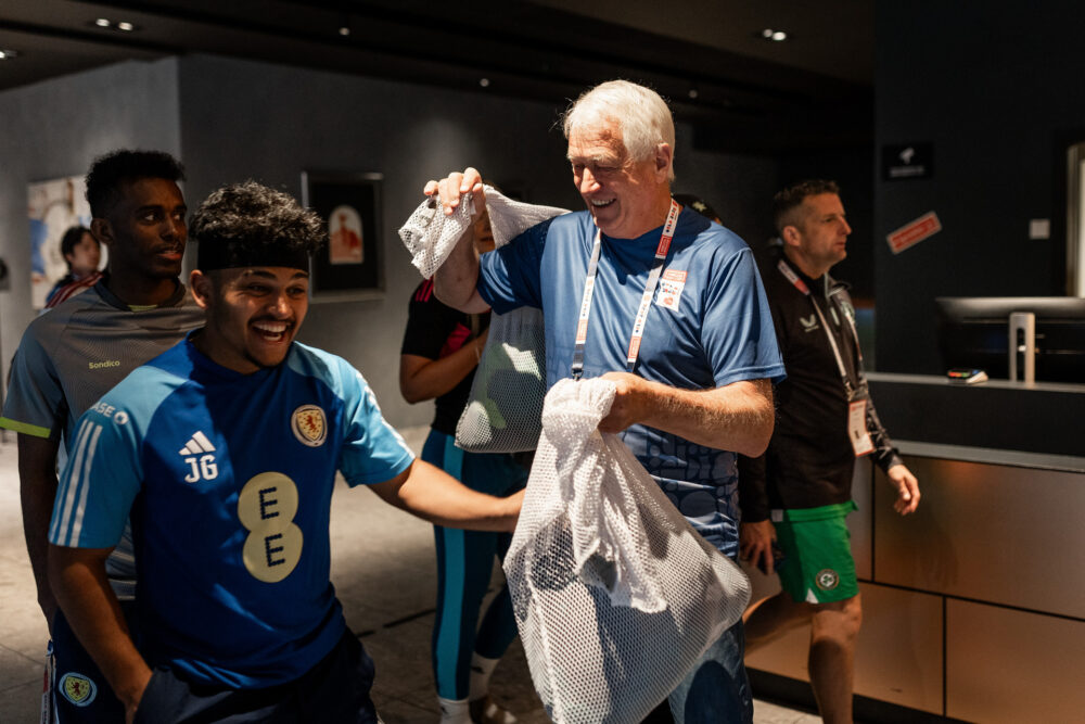 Foto: Steinar i blå hwc-t-skjorte smiler og gir en hvit sekk med klær til en spiller i blå t-skjorte som er på vei forbi ham. Spilleren smiler bredt. I bakgrunnen sees to andre spillere.