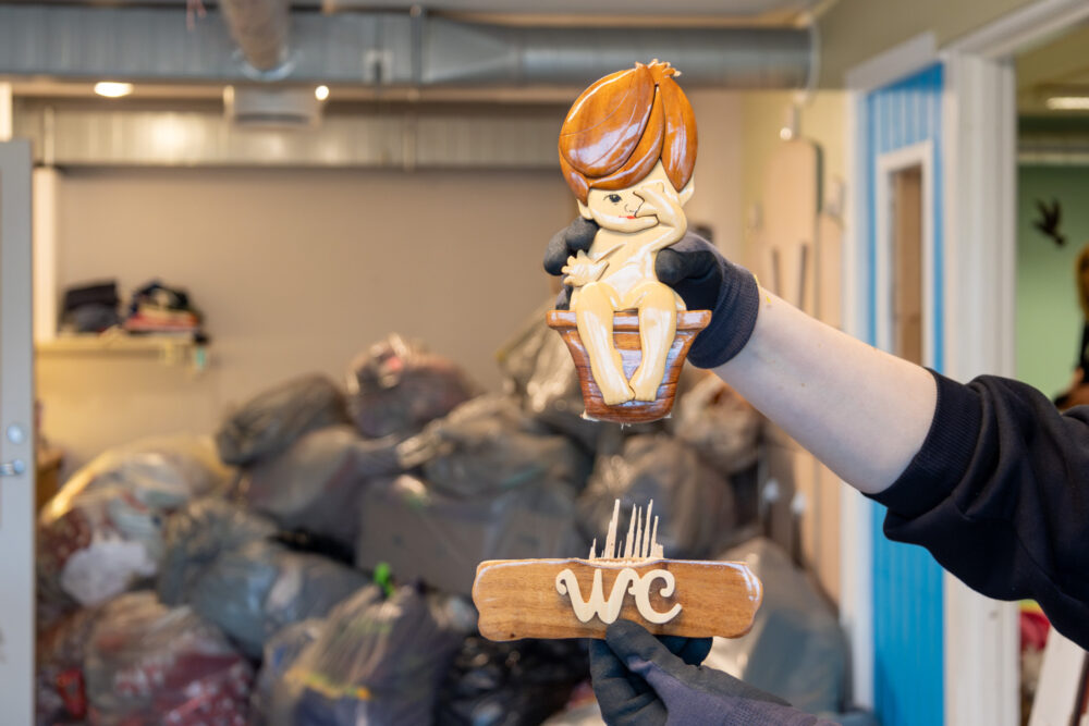 Foto: Arm med hanske på hånden holder opp en treskilt i to deler. På den øverste delen sitter en liten, naken gutt på potte. På den nederste delen står det WC,