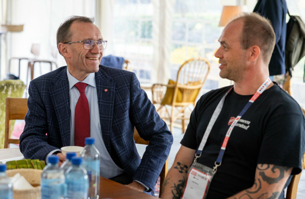 Foto: Mann med briller i mørk dress og rødt slips snakker til en mann i T-skjorte med tatoveringer. De sitter inne i et telt.