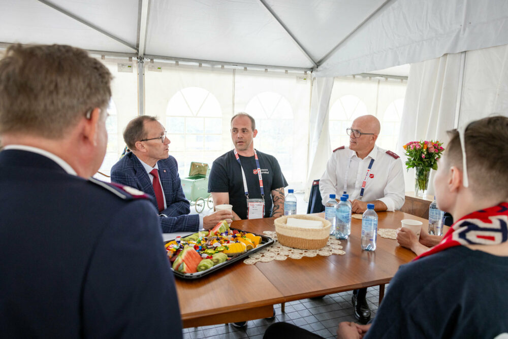 Foto: Fem personer sitter rundt et bord i et telt. To av dem er fra Frelsesarmeen og har på seg uniform. To er gatefotballspillere og siste er en mann med briller i mørkeblå dress med rødt slips.