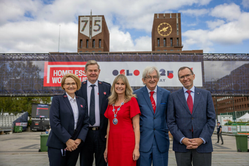 Foto: To kvinner og tre menn står pent kledde. I bakgrunnen Oslo rådhus med klokka og et banner i rødt, hvitt og blått med logoen til Homeless World Cup og teksten Oslo 2025.