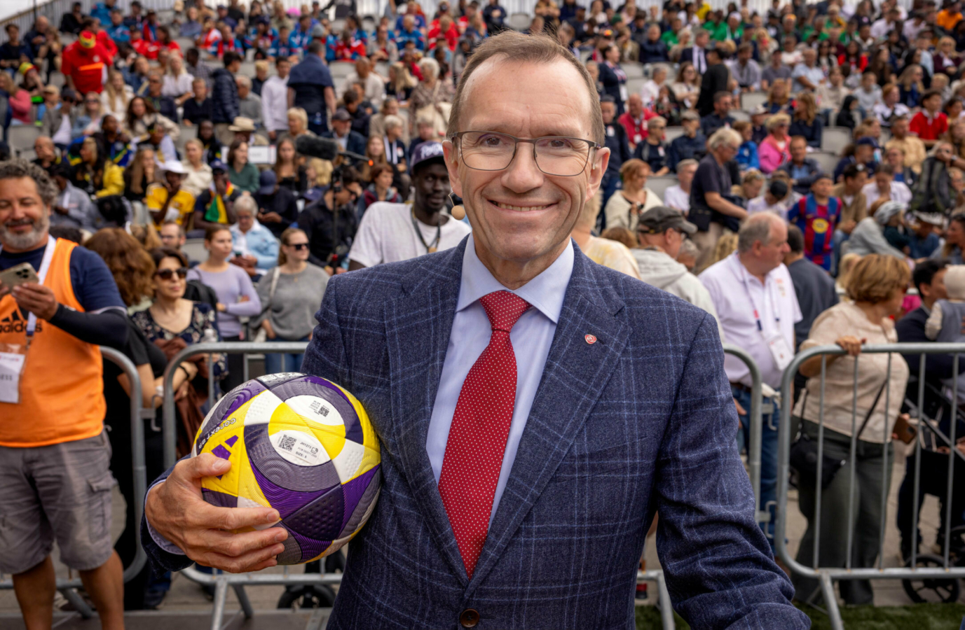Foto: Mann i blå dress med rødt slips holder en fotball. Han har mørkt hår, litt tynt på toppen, og briller. Bak ham er en tribune full av folk.