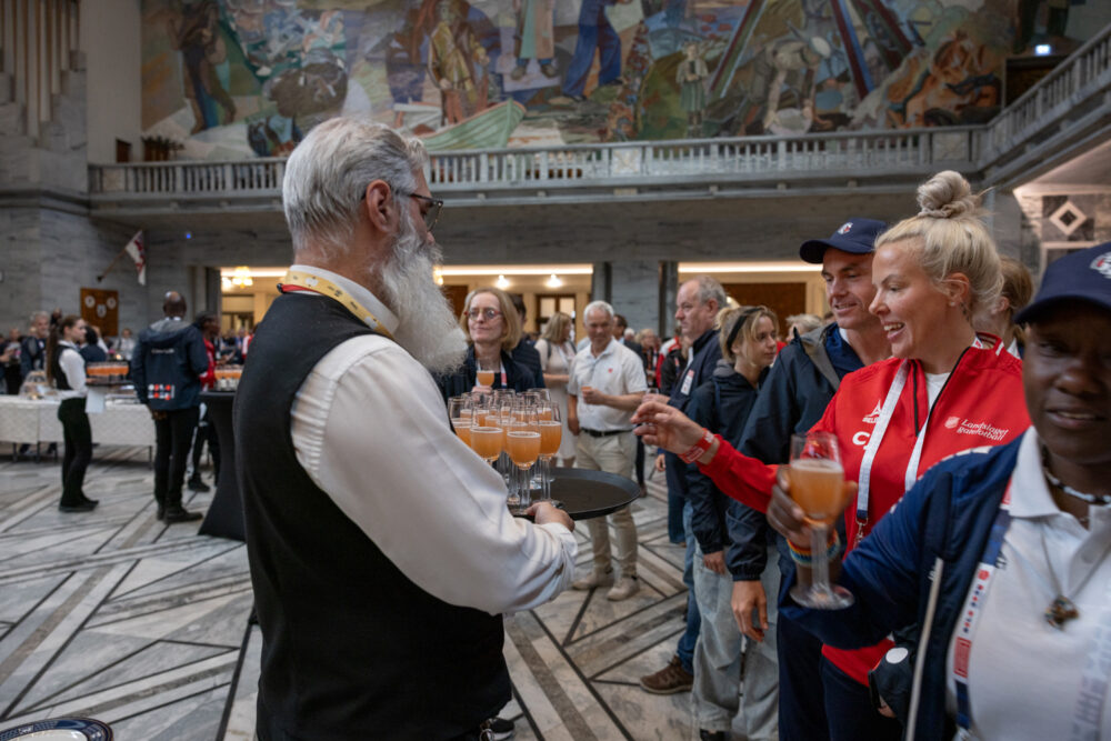 Foto: Kelner i hvit skjorte og svart vest med grått skjegg, serverer eple og rabarbra-drikke i glass med stett for en rekke folk i idrettsklær. En norsk jente med blondt, oppsatt hår og rød jakke med hvite striper forsyner seg av brettet.