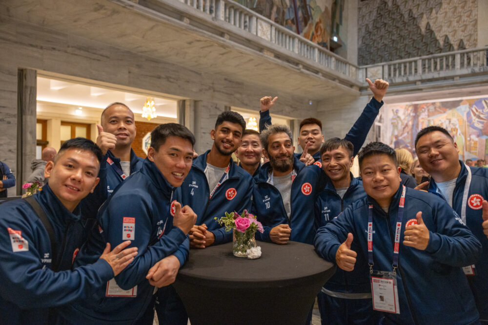 Foto: En gjeng mannlige spillere i blå drakter står rundt et høyt bord. Alle har svart hår, smiler og er glade. Flere viser tommel opp. En av dem strekker hendene i været.