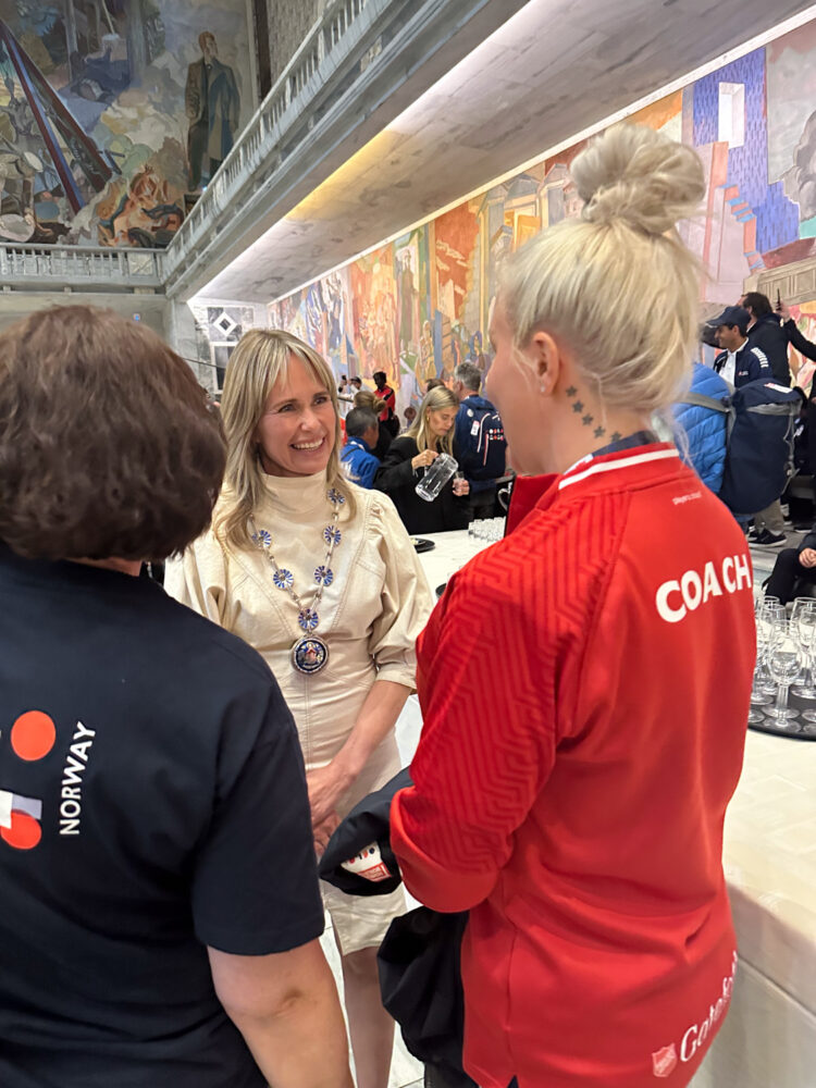 Foto: Blond kvinne i hvit lys kjole snakker med blond kvinne med oppsatt hår og rød treningsjakke. Ved siden av står en mørk kvinne med ryggen til i marineblå T-skjorte.