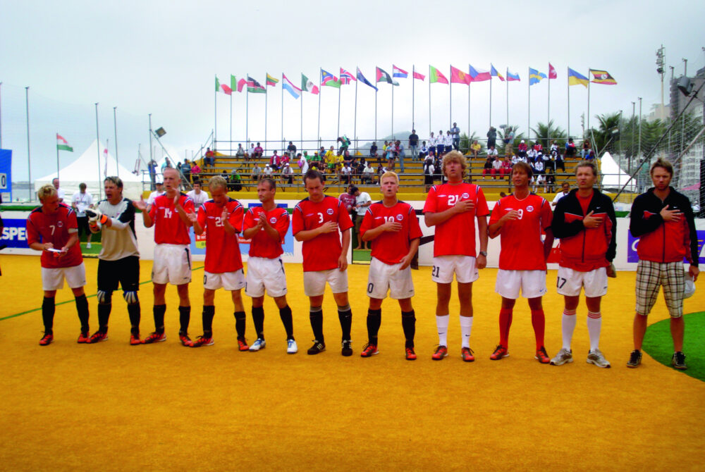 Det norske herrelandslaget i gatefotball oppstilt på en fotballbane, mens de synger nasjonalsangen med hånden på brystet.