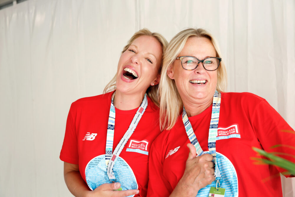 Foto: To blonde kvinner kvinner i røde fotballtrøyer lener seg mot hverandre og ler godt. På brystet står det Homeless World Cup.