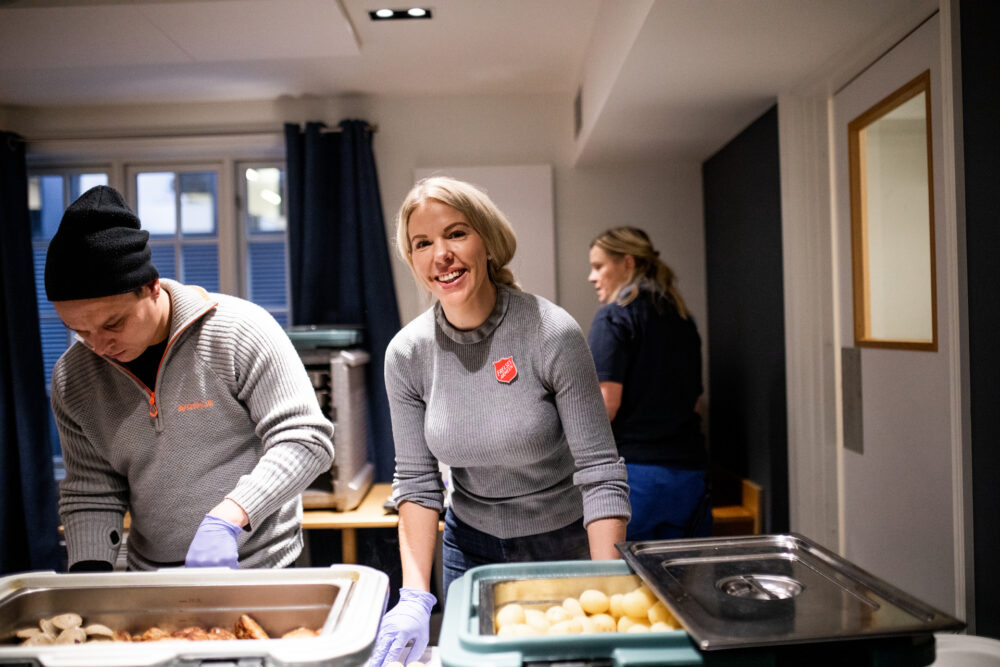 Kvinne og mann i Frelsesarmeenklær jobber smilende med matservering innendørs.