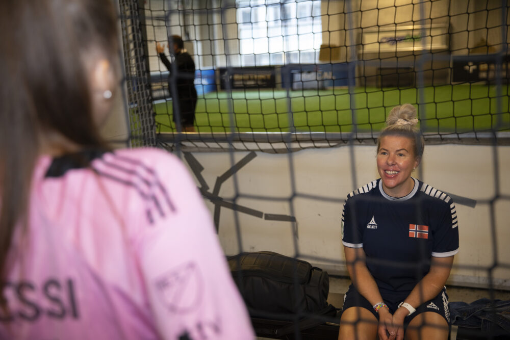 To kvinner i samtale ved en innendørs fotballbane. Cathrine smiler mens hun hviler på en benk.