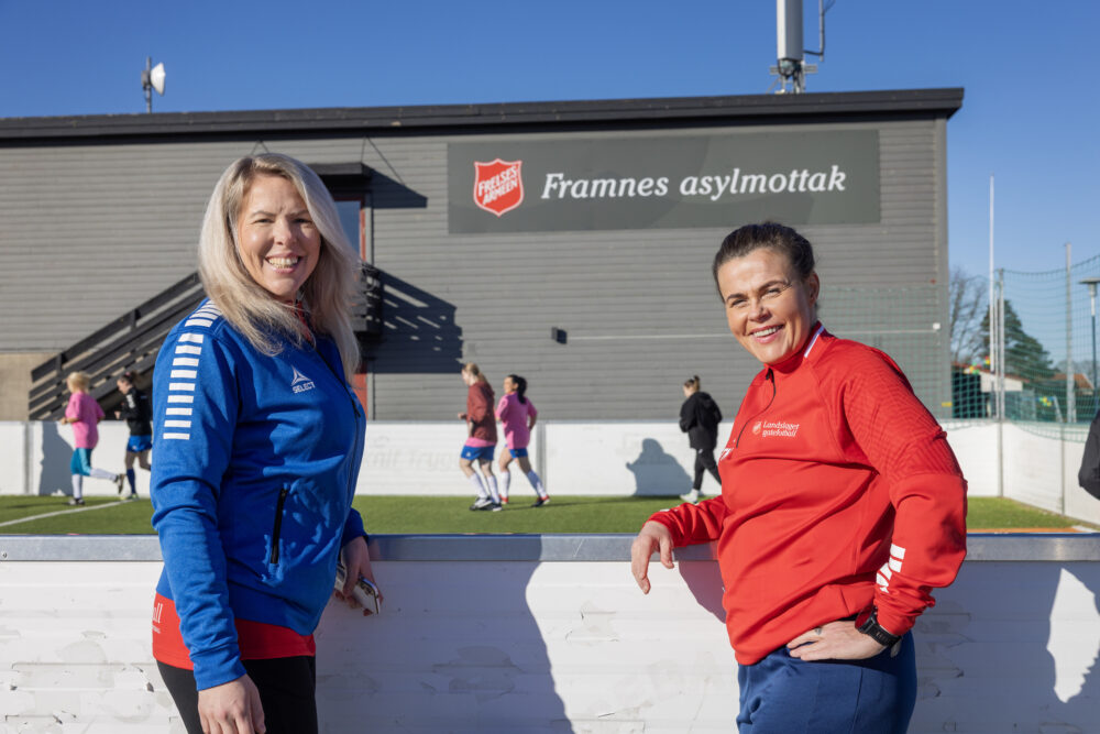 To kvinner står smilende foran en fotballbinge hvor en gruppe jenter spiller fotball.