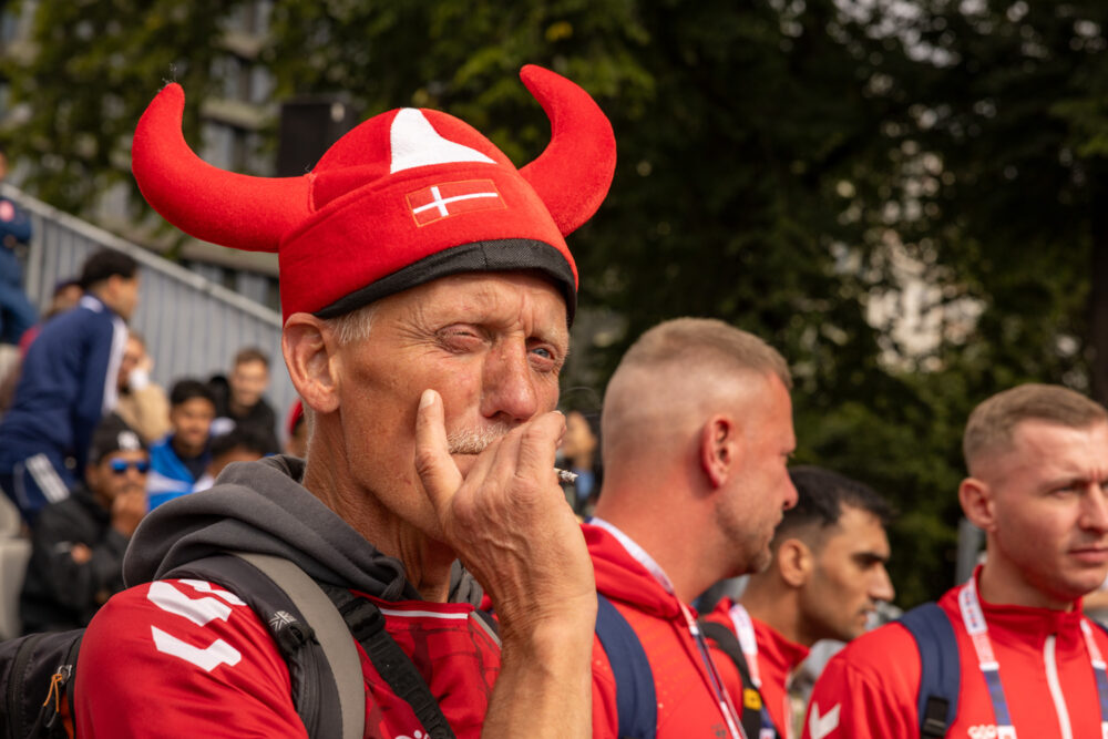Foto: Eldre mann med grå bart under en rød vikinghjelm i stoff. Han røyker en sigarett. Bak ser man folk på tribunen.