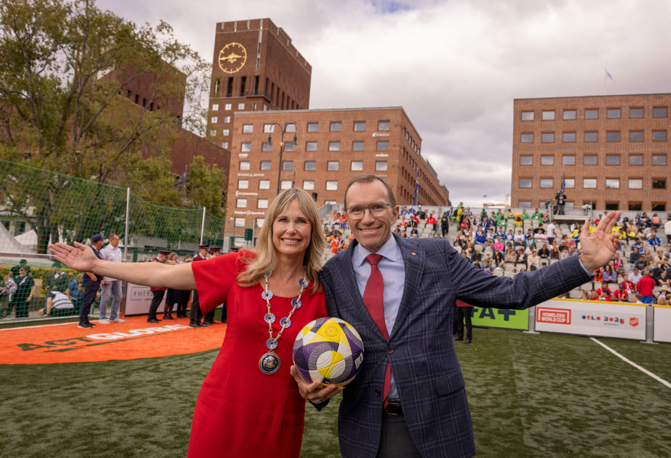 Foto: Kvinne i rød kjole med blondt hår og ordførerkjede står tett sammen med en mann i mørk dress og rødt slips. Han er mørk i håret og har briller. De strekker ut hver sin arm i en velkomst. I bakgrunnen sees Oslo Rådhus i rød murstein.