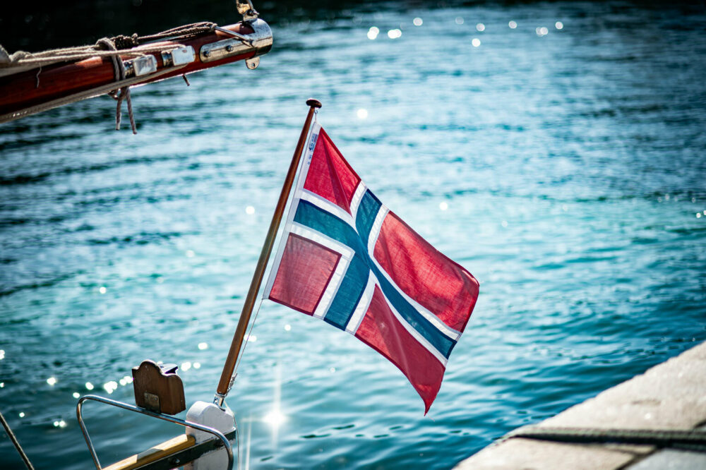 Foto: Norsk flagg bak på en seilbåt. Det glitrer i vannet.