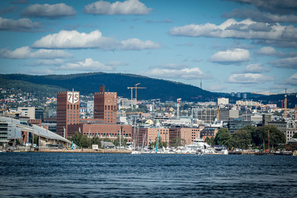 Foto: Tatt fra en båt der det er vann foran. Bak ligger byen og en stor, rød bygning ruver i front.