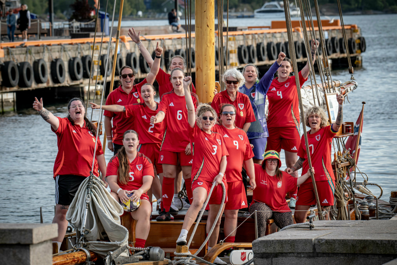 Foto: Et kvinnelag i røde fotballdrakter vinker fra en seilbåt som ligger til kai.