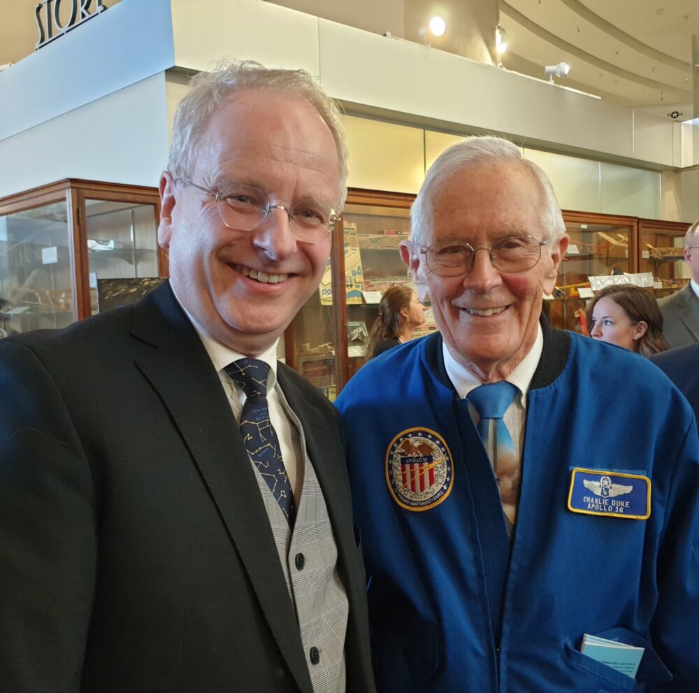 Foto: En norsk mann i slutten av 50-årene står sammen med en pensjonist i en knall blå jakke med merke fra Apollo 16 og et annet pilotmerke. Begge har briller og grått hår.