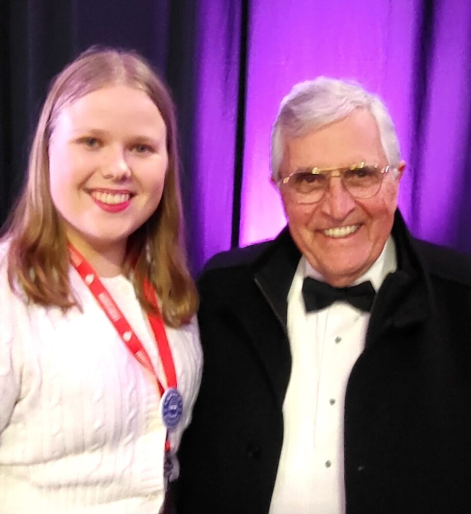 Foto: Ung jente i hvit genser står sammen med en pensjonert mann med svart dressjakke, hvit skjorte og sløyfe. Han har briller. I bakgrunnen er det et litt sceneteppe.