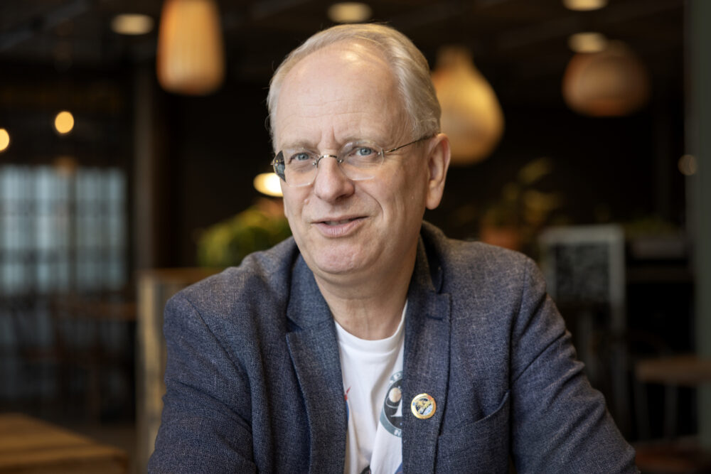 Foto: Mann i blå dressjakke med briller sitter i et større rom.