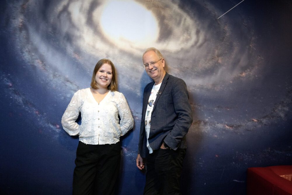 Foto: Mann med briller og dressjakke sammen med ung jente med hvit bluse. De står mot en vegg som har et bilde av verdensrommet.