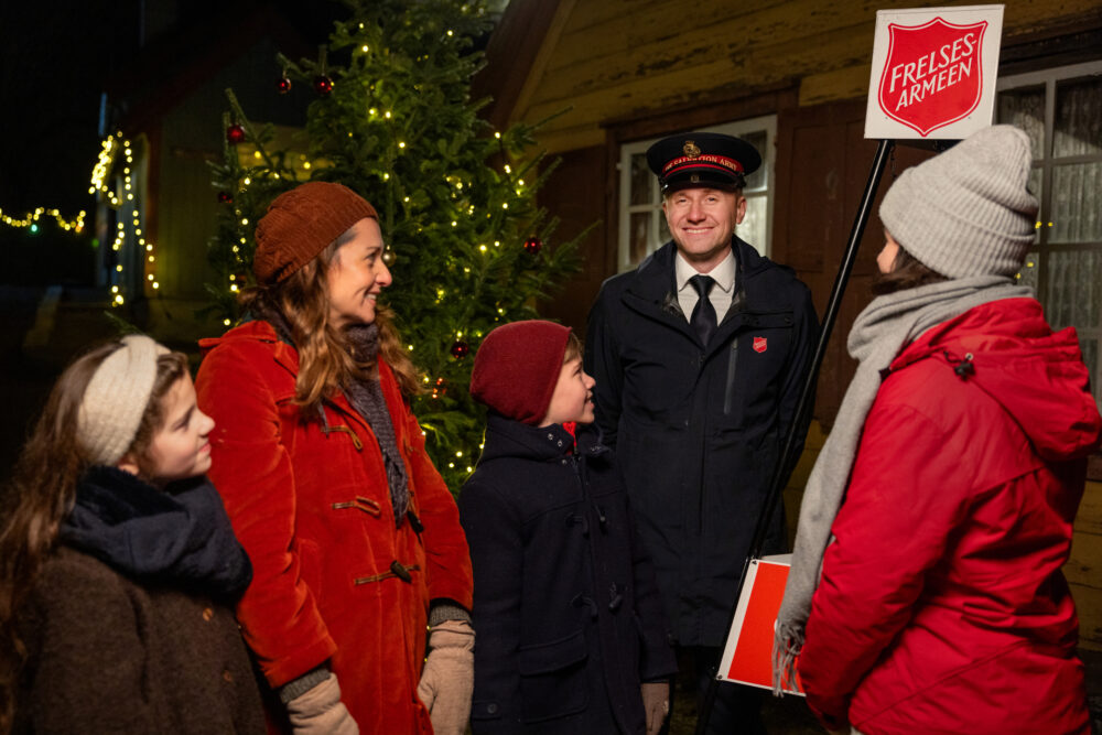 Foto: Ung mann i uniform fra Frelsesarmeen står ute ved en julegryte. Rundt ham står to kvinner og to barn. De har på seg vintertøy og i bakgrunnen er det juletre og julelys.