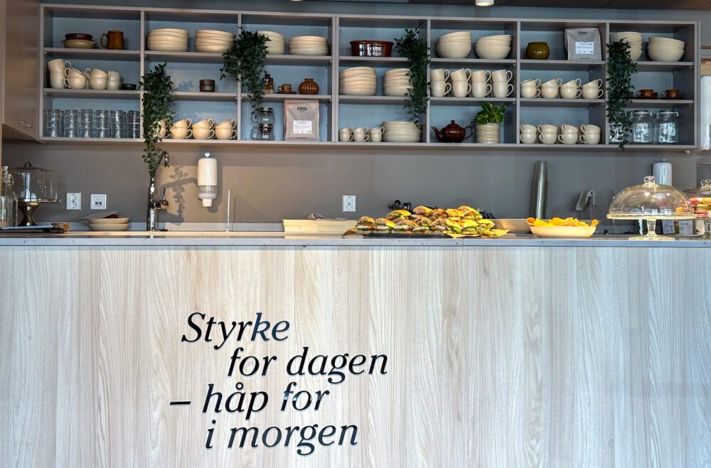 Foto: Av kafédisken med alle hyller bak, fulle av kopper, glass og skåler. Foran på det lyse treverket i disken står det en tekst.