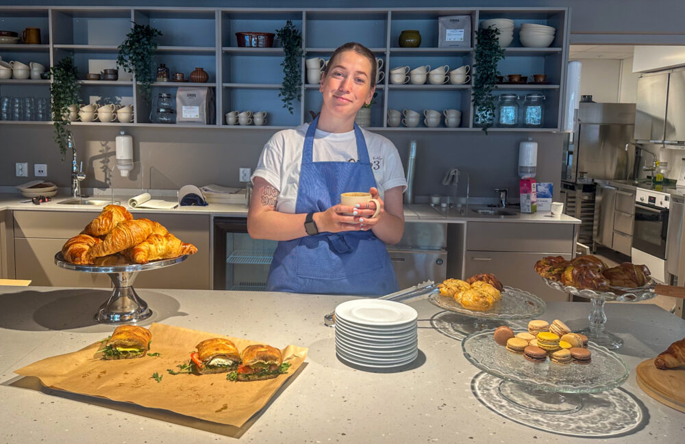 Foto: Kvinne holder i en kaffekopp. Hun står bak en kafédisk og har på seg hvit T-skjorte og blått forkle.