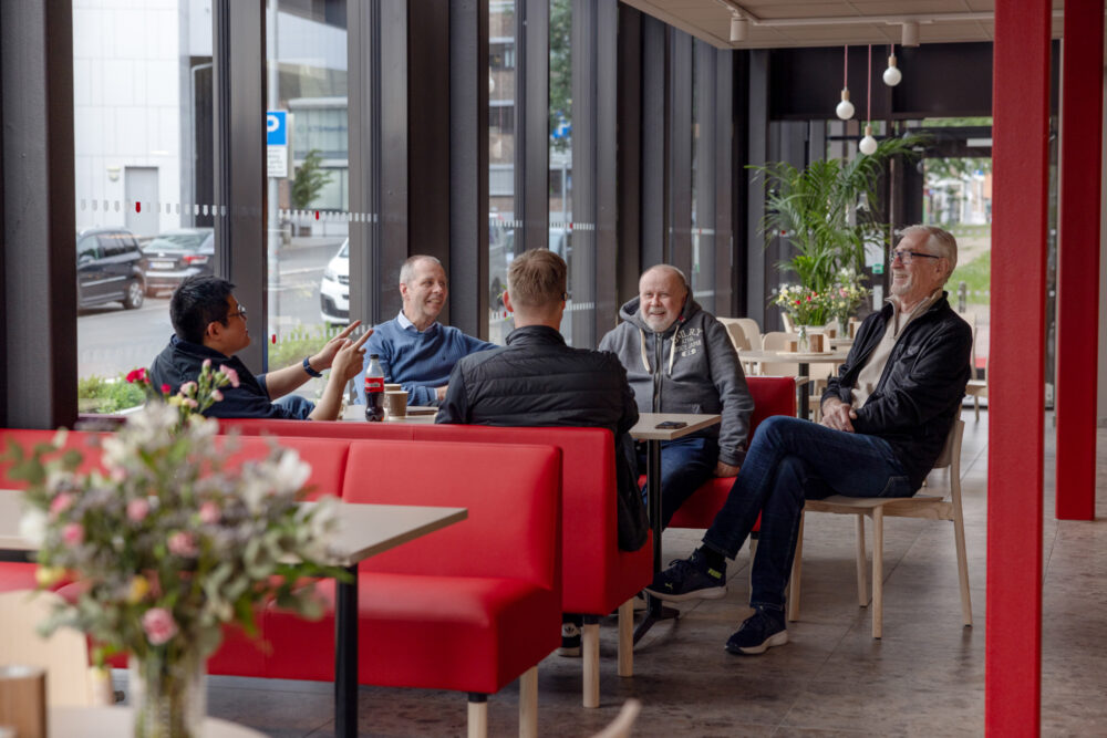 Foto: Fem voksne menn sitter i en sittegruppe. De prater og ler.