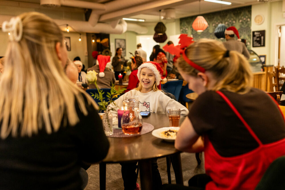 Foto: Ung jente ed nisselue sitter på et bord og snakker med to eldre jenter med ryggen til. Lokalet er fullt av folk.