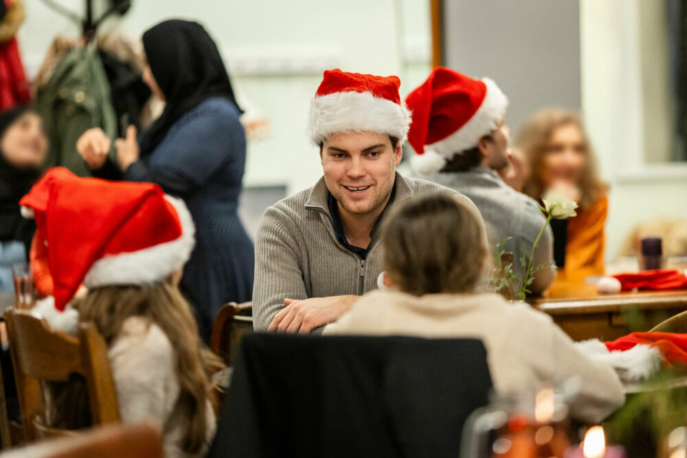 Foto: Ung mann i grå genser sitter med nisselue på og snakker med noen barn i småskolealder. De sitter med ryggen til. Han smiler og har mørkt hår.