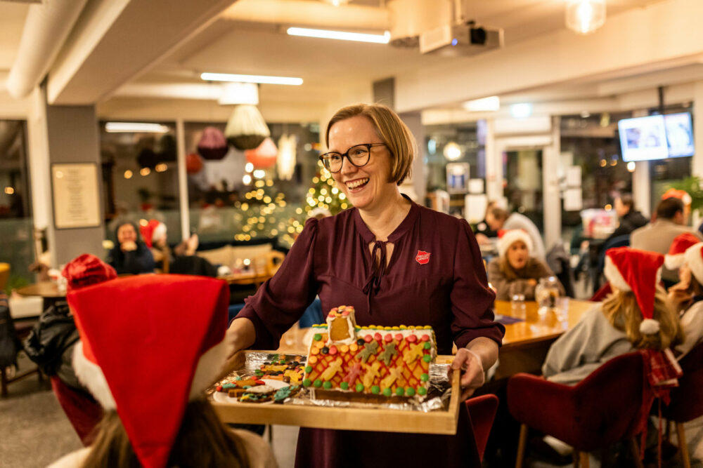Foto: Kvinne tidlig i 40-årene med burgunder penkjole og logo fra Frelsesarmeen. Hun er blond og har briller. I hendene bærer bærer hun et brett med et pyntet pepperkakehus. Det er folk på alle kanter i rommet.
