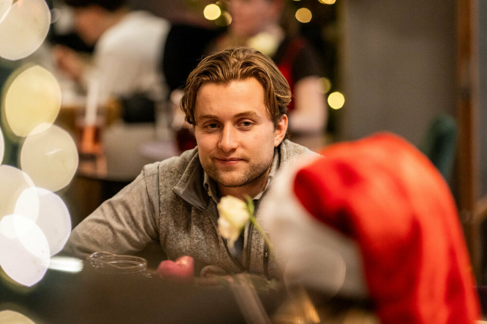 Foto: Ung mann med mørkeblondt hår siter ved et bord og snakker til et barn med nisselue.