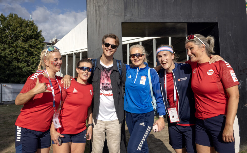 Foto: Kvinner i røde og blå drakter står på hver sin side av en høy mann med solbriller.