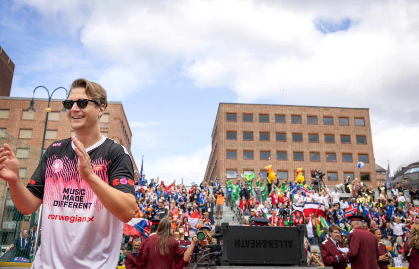 Foto: Mann med solbriller og fotballtrøye skal til å klappe i hendene. På tribunen bak er det masse folk med flagg.