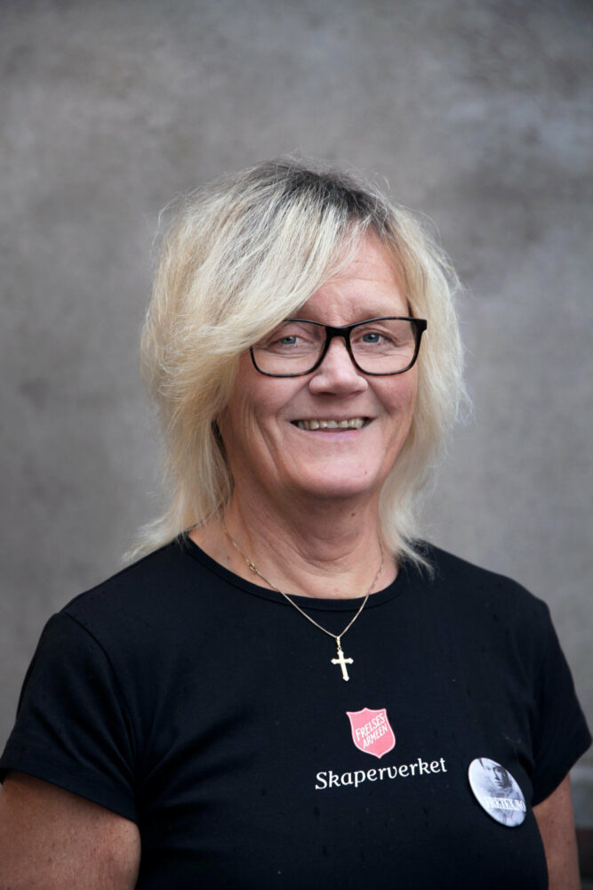 Foto: Portrett av kvinne i 50-årene med gråblondt hår og briller. Hun har på seg en svart T-skjorte med den røde logoen til Frelsesarmeen på.