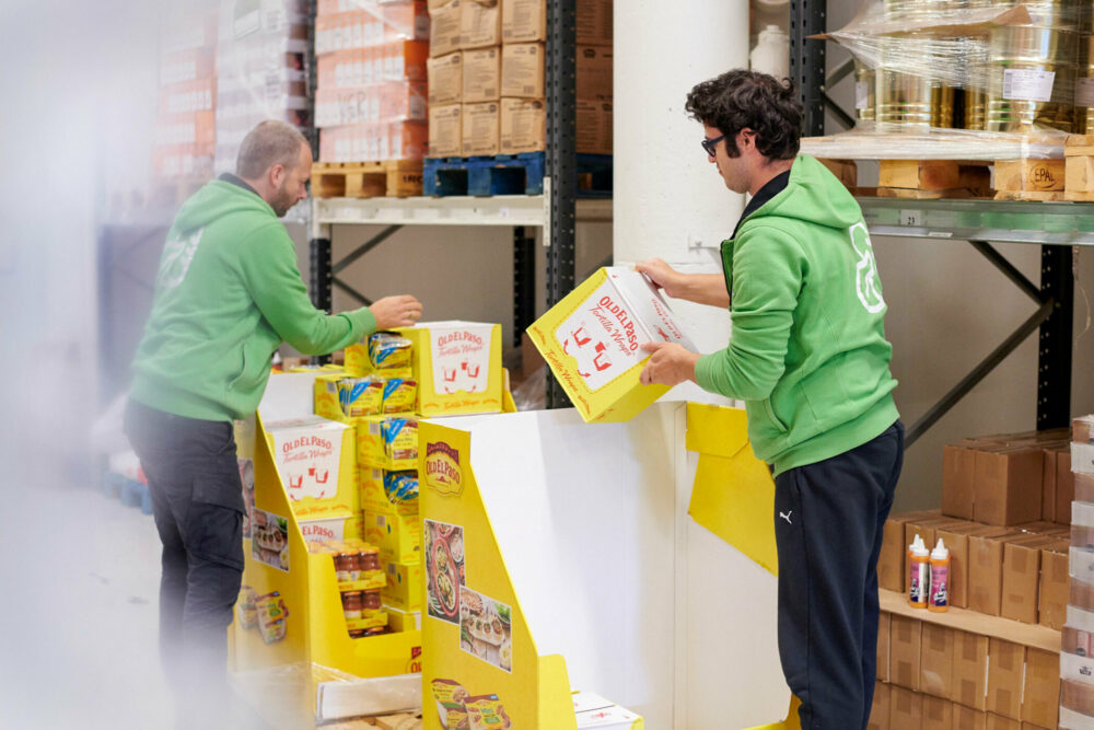 Foto: To mennesker i grønne klær løfter opp esker med mat fra en pall. I bakgrunnen ser vi høye hyller fylt av matvarer.