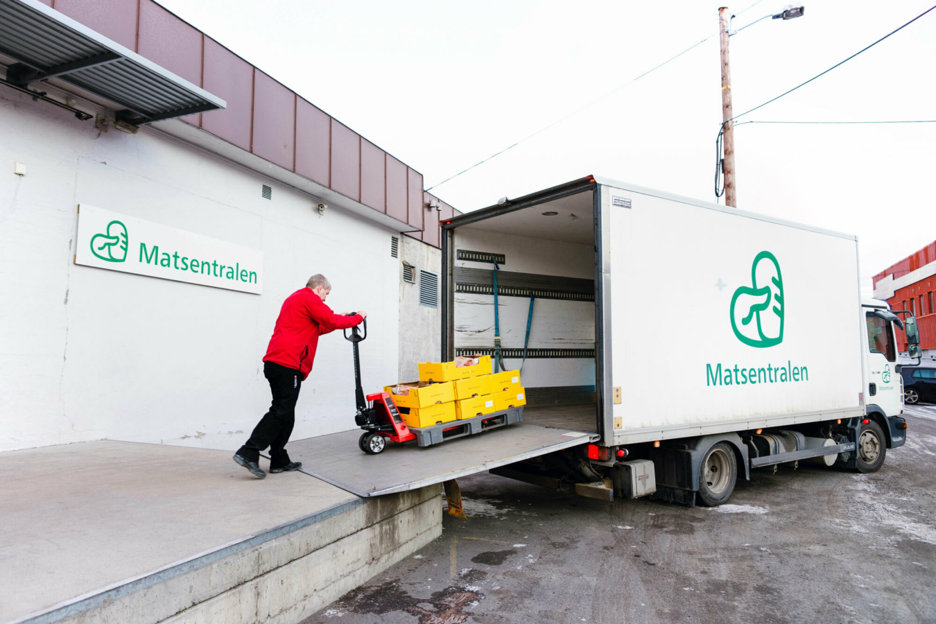 Foto: En mann i rød flisjakke triller en pall på en stor, hvit varebil. På siden av bilen, og på bygget det losses fra, står logoen til Matsentralen.