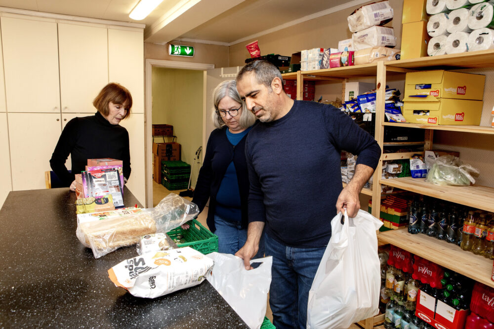 Foto: Bildet er tatt i rommet der matposene pakkes. Margrete, Gerd Britt og Fatih står ved et bord mitt i rommet, hvor det ligger ulike matvarer. Fatih holder to ferdigpakkede poser. Bak dem er store hyller med tørrmat og toalettpapir.