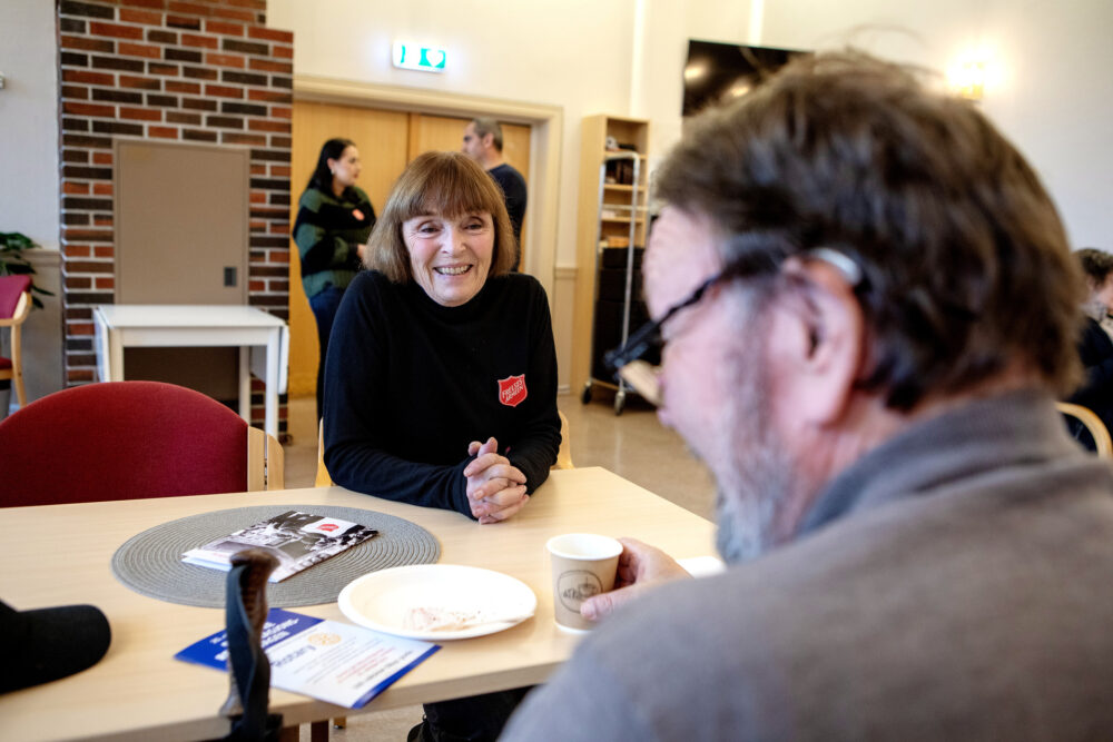 Foto: Eldre kvinne med mørkt hår sitter ved et bord og snakker med en mann som sitter med ryggen til. Hun har på seg blå genser med logo fra Frelsesarmeen.