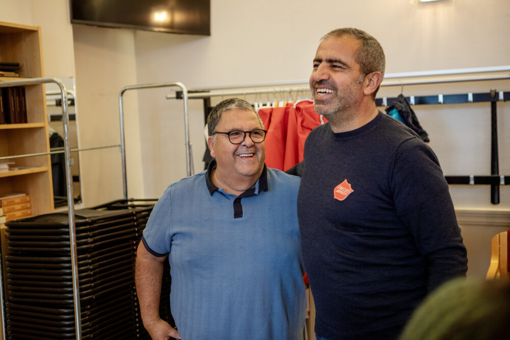 Guillermo i lys blå t-skjorte står og smiler ved siden av Fatih i svart genser. Bak dem er et stativ med en stor stabel sammenslåtte plaststoler