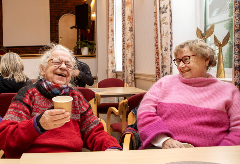 Foto: To eldre damer sitter ved siden av hverandre ved et bord. Charlott i rød strikkegenser holder en kaffekopp, hun lener seg tilbake og ler. Elfi i rosa strikket genser ser på henne og smiler.