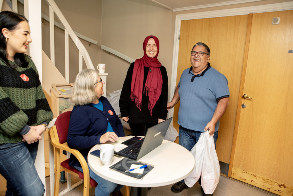 Foto: Middelaldrende kvinne med grått hår sitter foran en pc ved et lite bord. Hun har logo fra Frelsesarmeen på den blå strikkejakken. Bak er en hvit trapp opp til etasjen over. Rundt henne står to kvinner og en mann. Han bærer noen hvite poser.