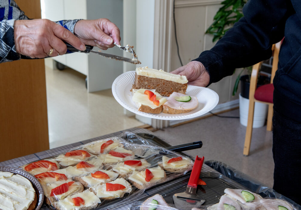 Foto: Nærbilde av en tallerken med to brødskiver og et kakestykke. Bare hendene til den som holder tallerkenen og til den frivillige som legger på maten synes, Et stort fat med brødskiver med ost sees på bordet under.