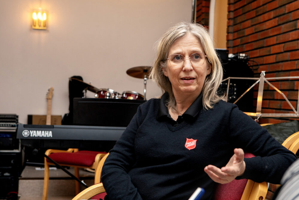 Foto: Kvinne i 50-årene med briller og gråblondt hår sitter i en sofa. Hun gestikulerer med den ene hånden når hun snakker. På den blå genseren er en rød logo fra Frelsesarmeen.