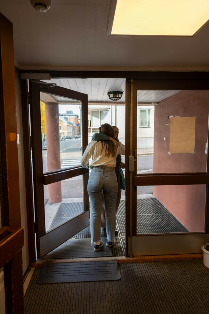 Foto: Dhurata gir Inese en god klem i døra på vei ut fra Frelsesarmeens lokaler. En gate og et hvitt hus sees i bakgrunnen. Dhurata står med ryggen til kamera, Inese sees så vidt bak henne.
