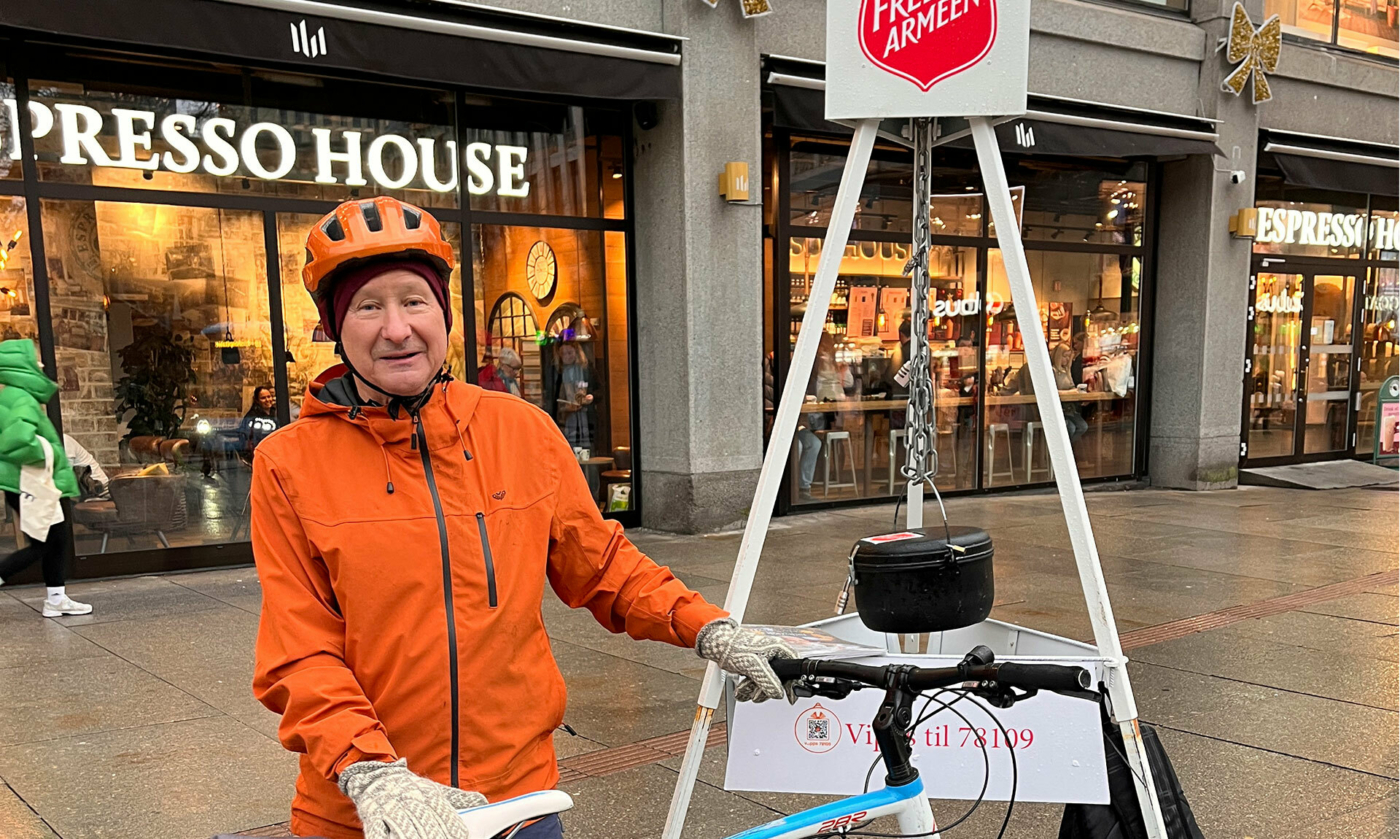 Foto: Eldre mann i oransje jakke og hjelp holder i en sykkel. Han står foran et stativ med Frelsesarmeens julegryte hengende ned i en kjetting. Han står foran det som er et kjøpesenter med kafé på første plan.