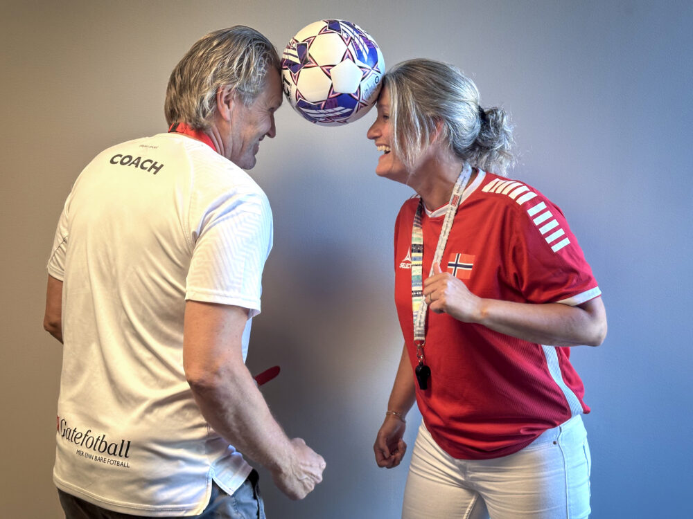 Foto: Mann og kvinne i hvit og rød fotballtrøye holder en ball mellom hodene sine. Begge ler.