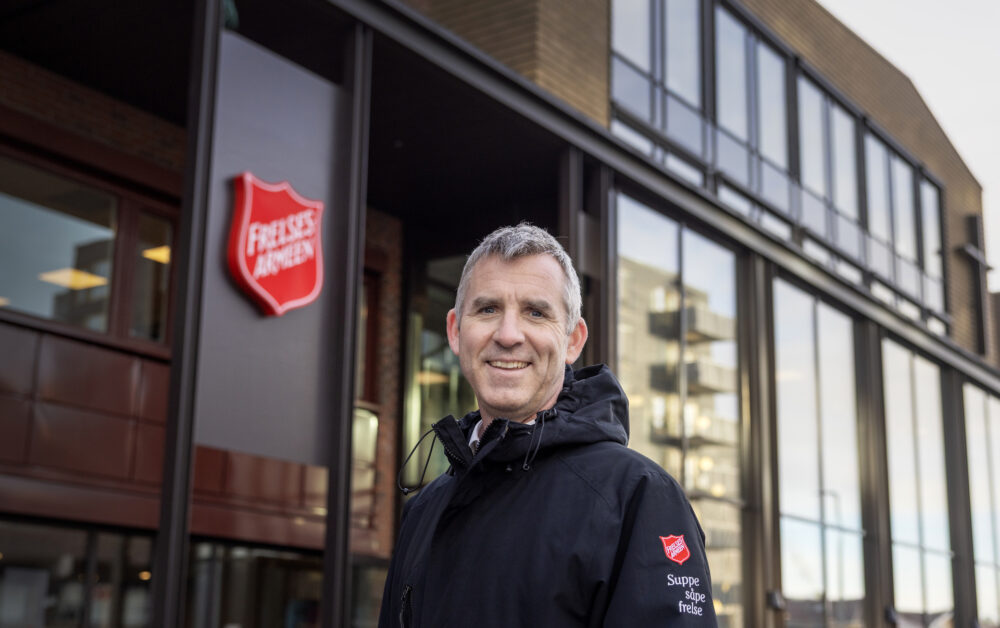 Foto: Smilende mann i 50-årene med grålig hår står utenfor et bygg med en stor logo fra Frelsesarmeen. Han har på seg en mørk jakke med logo fra Frelsesarmeen på armen. Under står det i hvit skrift står det: Suppe, såpe, frelse.