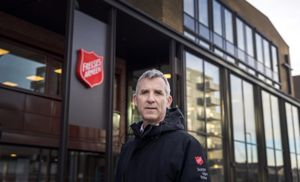 Foto: Mann i 50-årene med grålig hår står utenfor et bygg med en stor logo fra Frelsesarmeen. Han har på seg en mørk jakke med logo fra Frelsesarmeen på armen. Under står det i hvit skrift står det: Suppe, såpe, frelse.
