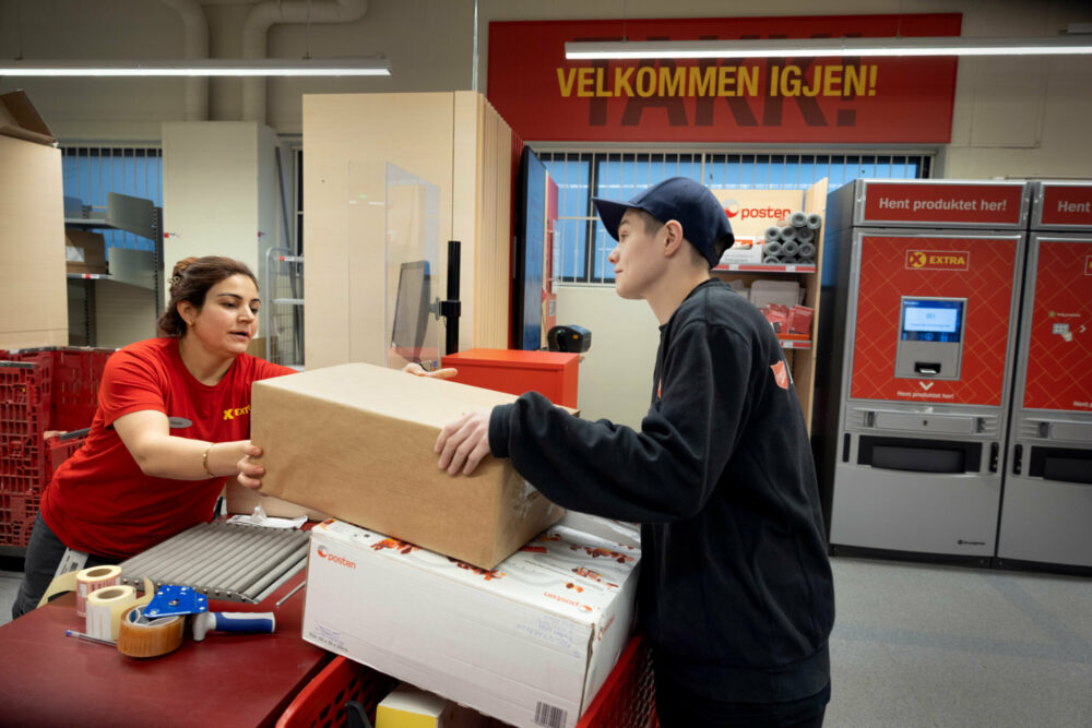 Foto: En dame bak skranken i posten leverer to store pakker til en ung dame med blå caps og kort hår.