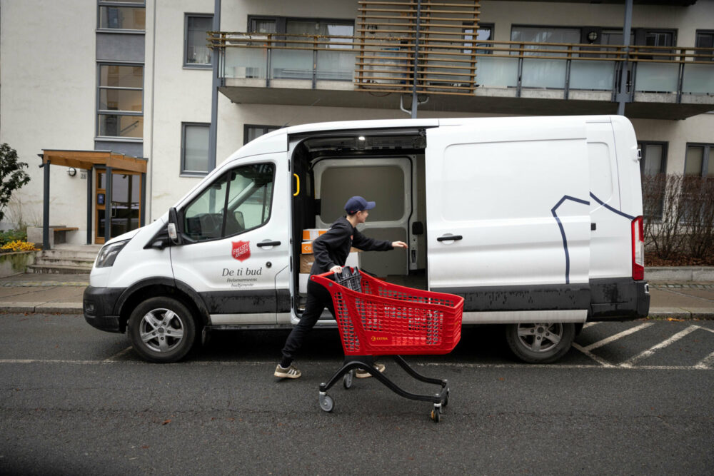 Foto: Kvinne med rød handlevogn utenfor en hvit varebil fra Frelsesarmeen. Den står parkert utenfor en moderne blokk.